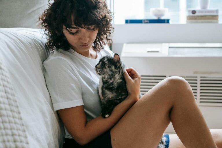 cat realationship: side view of crop young female with curly hair in casual clothes hugging and stroking adorable purebred cat while relaxing at home
