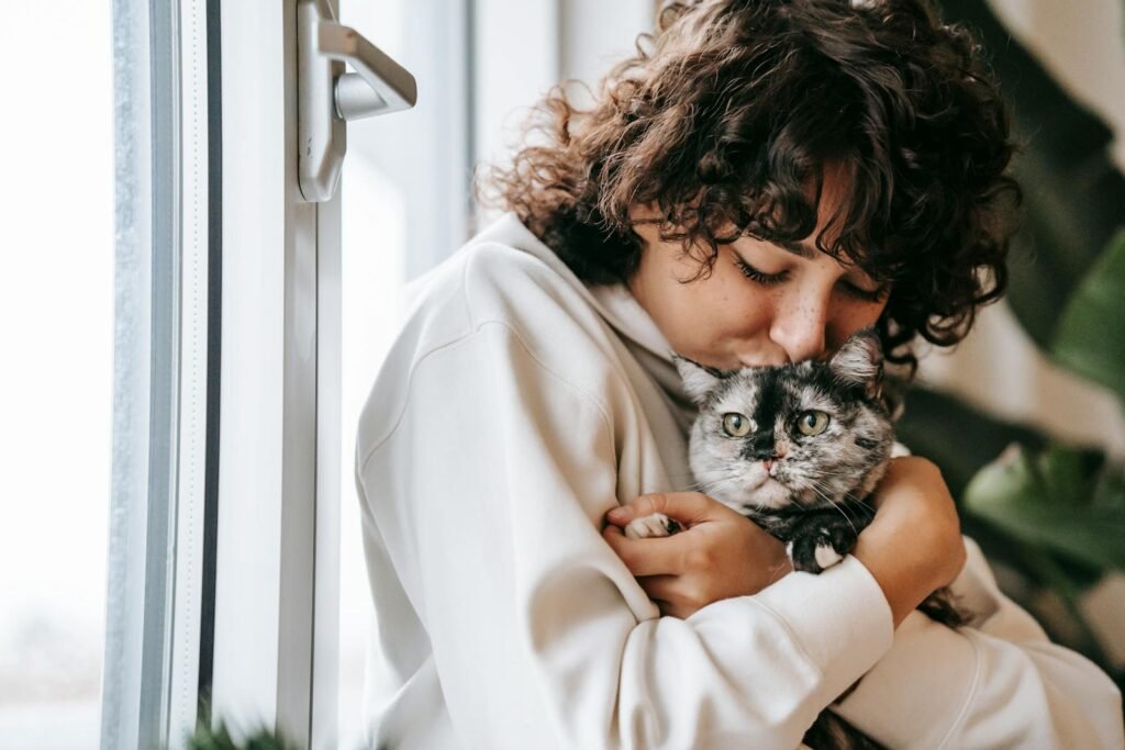 A woman affectionately hugs her cat by the window, creating a warm and tender moment indoors.