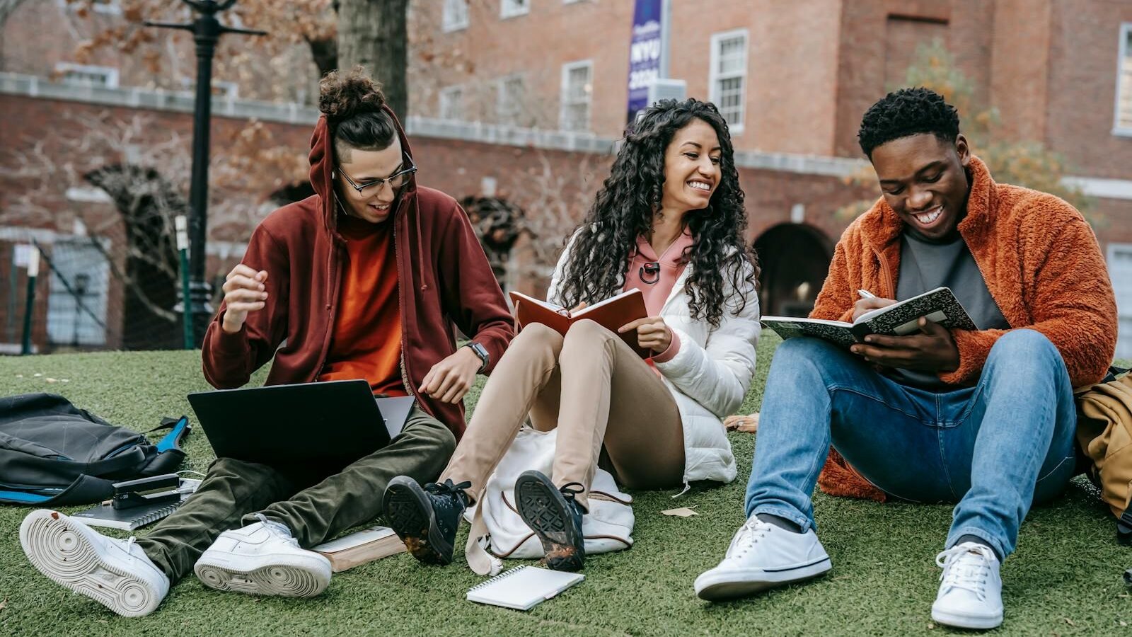 A group of three diverse students sitting on a grassy area, smiling and engaged in reading books and working on a laptop, with a university campus building in the background.