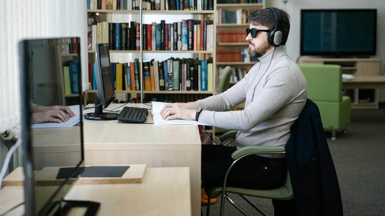 A man wearing headphones and sunglasses sits at a desk in a modern office, working on a computer with bookshelves in the background.