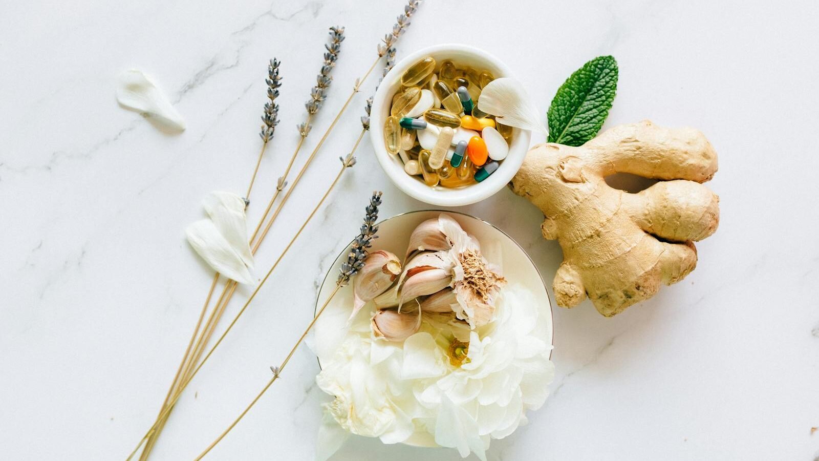 A collection of natural remedies and supplements, including ginger root, lavender, and a small bowl of mixed capsules, arranged on a marble surface.