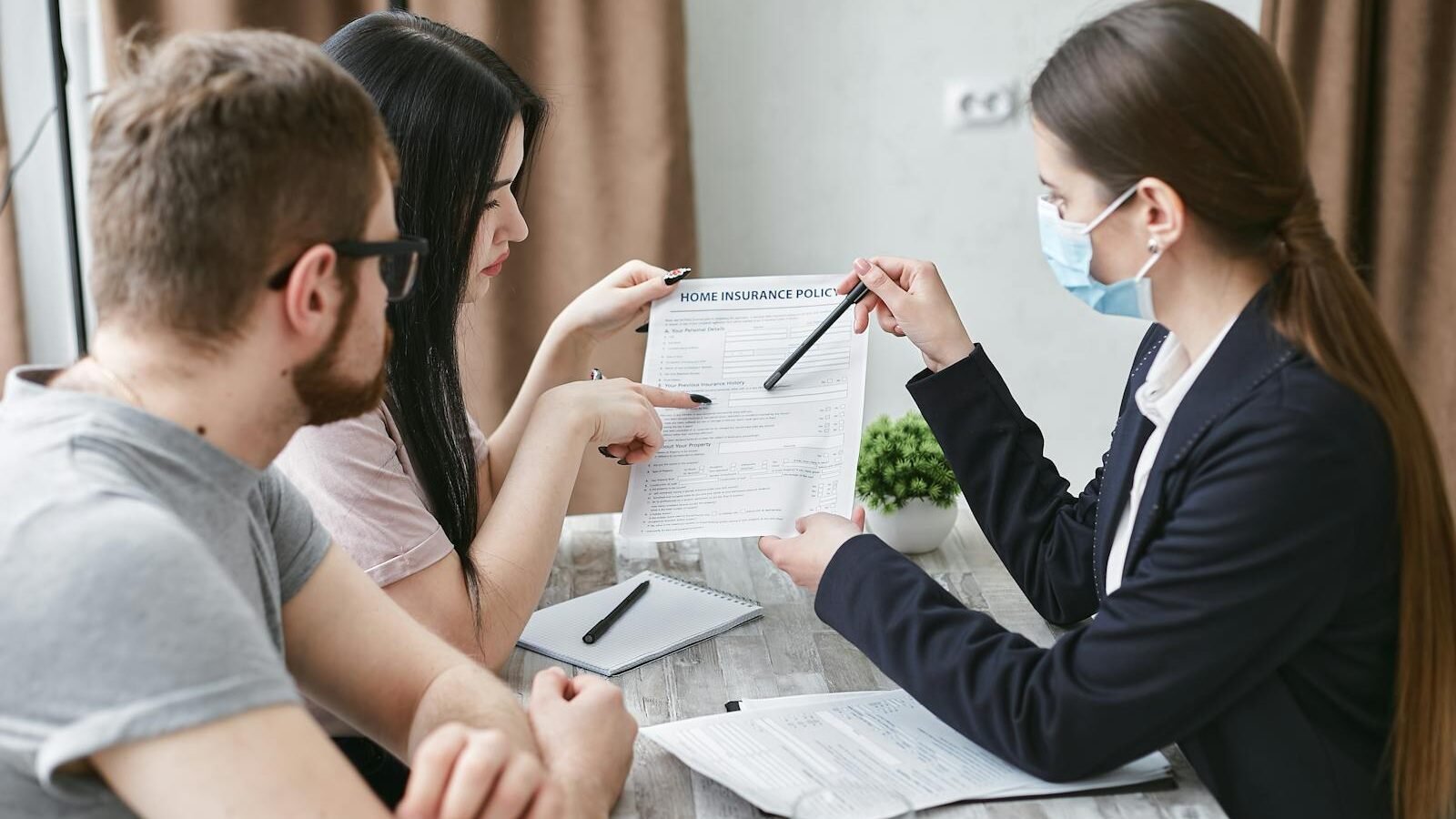 A professional wearing a face mask discussing documents with a couple at a table, pointing at details on the paperwork, in a well-lit indoor setting.
