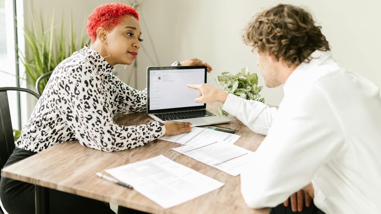 Two professionals engaged in a discussion at a desk, with one person pointing at a laptop screen while documents are spread out, showcasing collaboration and communication.