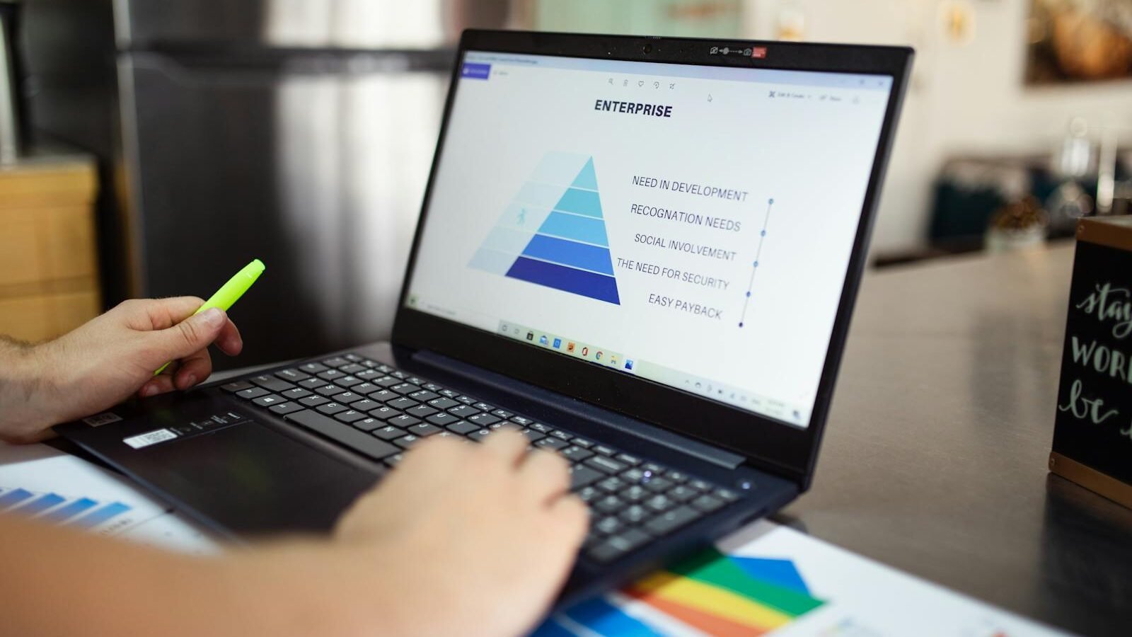 A person holding a highlighter while working on a laptop displaying a pyramid chart, with colorful data sheets spread on the table.