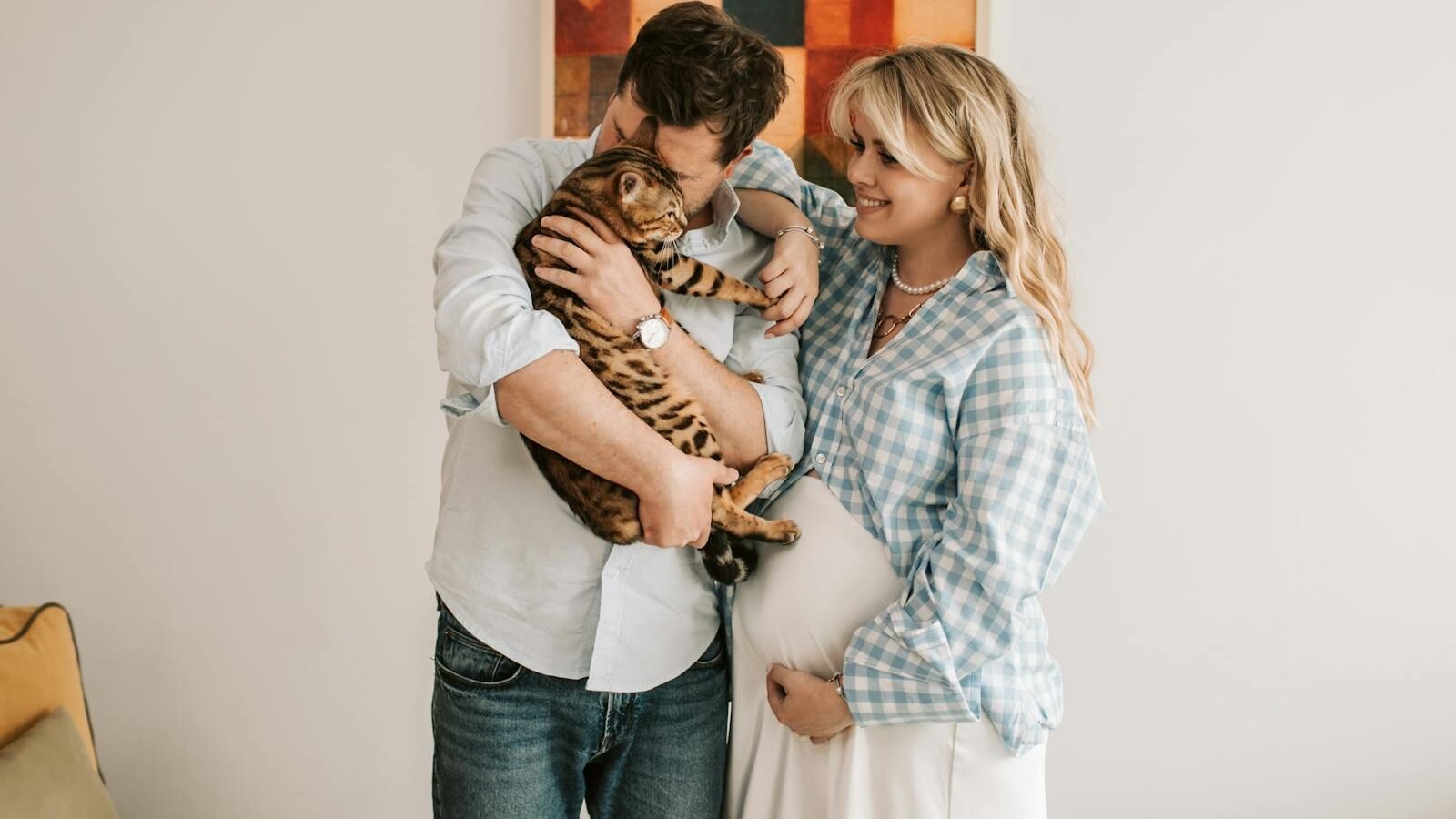 A happy couple, with the man holding and cuddling a Bengal cat and the woman, who is pregnant, smiling warmly beside them in a cozy home setting.