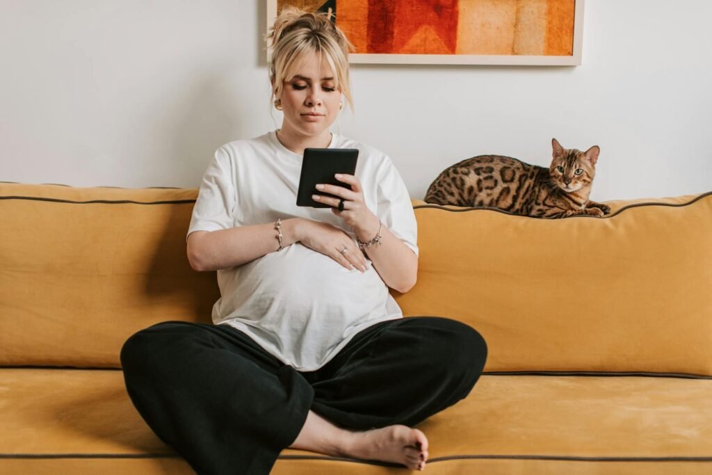 cat confusion: Pregnant woman relaxing on a sofa with a Bengal cat nearby, reading a tablet in a cozy indoor setting.