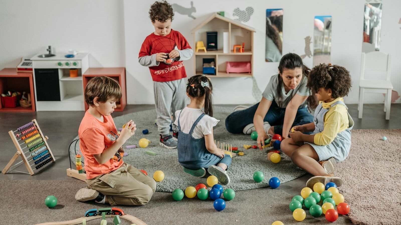 A group of young children playing with colorful toys in a preschool setting while a teacher interacts with them, promoting early socialization.