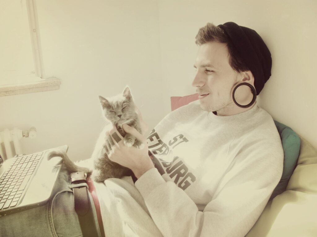 A man wearing a beanie and large ear gauges relaxes with a content gray cat on his lap, gently holding its chin while sitting near a laptop.
