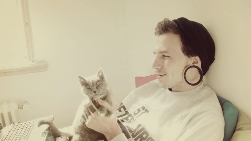 A man wearing a beanie and large ear gauges relaxes with a content gray cat on his lap, gently holding its chin while sitting near a laptop.