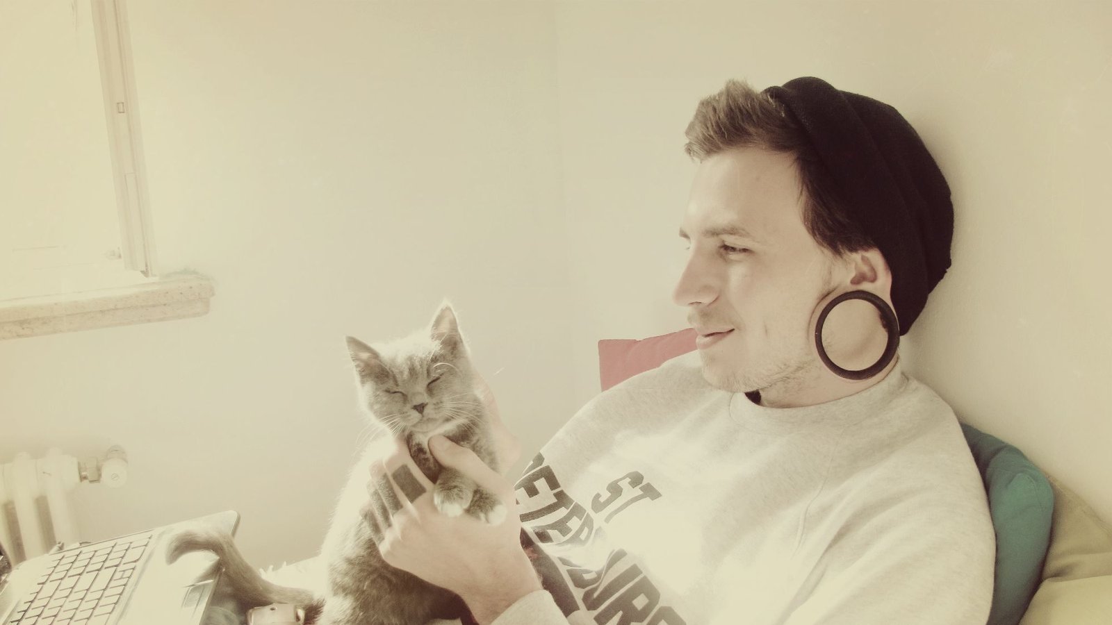 A man wearing a beanie and large ear gauges relaxes with a content gray cat on his lap, gently holding its chin while sitting near a laptop.