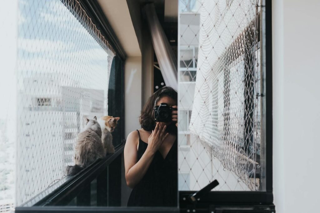 A woman taking a picture with a camera reflected in a window, with two cats on a balcony.
