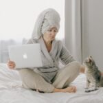 A woman sits comfortably on a bed with her laptop, wearing a bathrobe and towel, petting a cat.