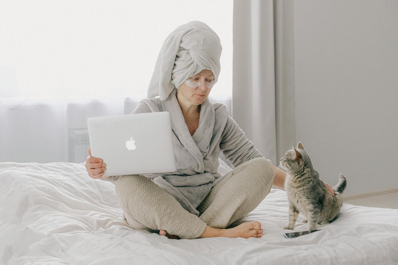 A woman sits comfortably on a bed with her laptop, wearing a bathrobe and towel, petting a cat.