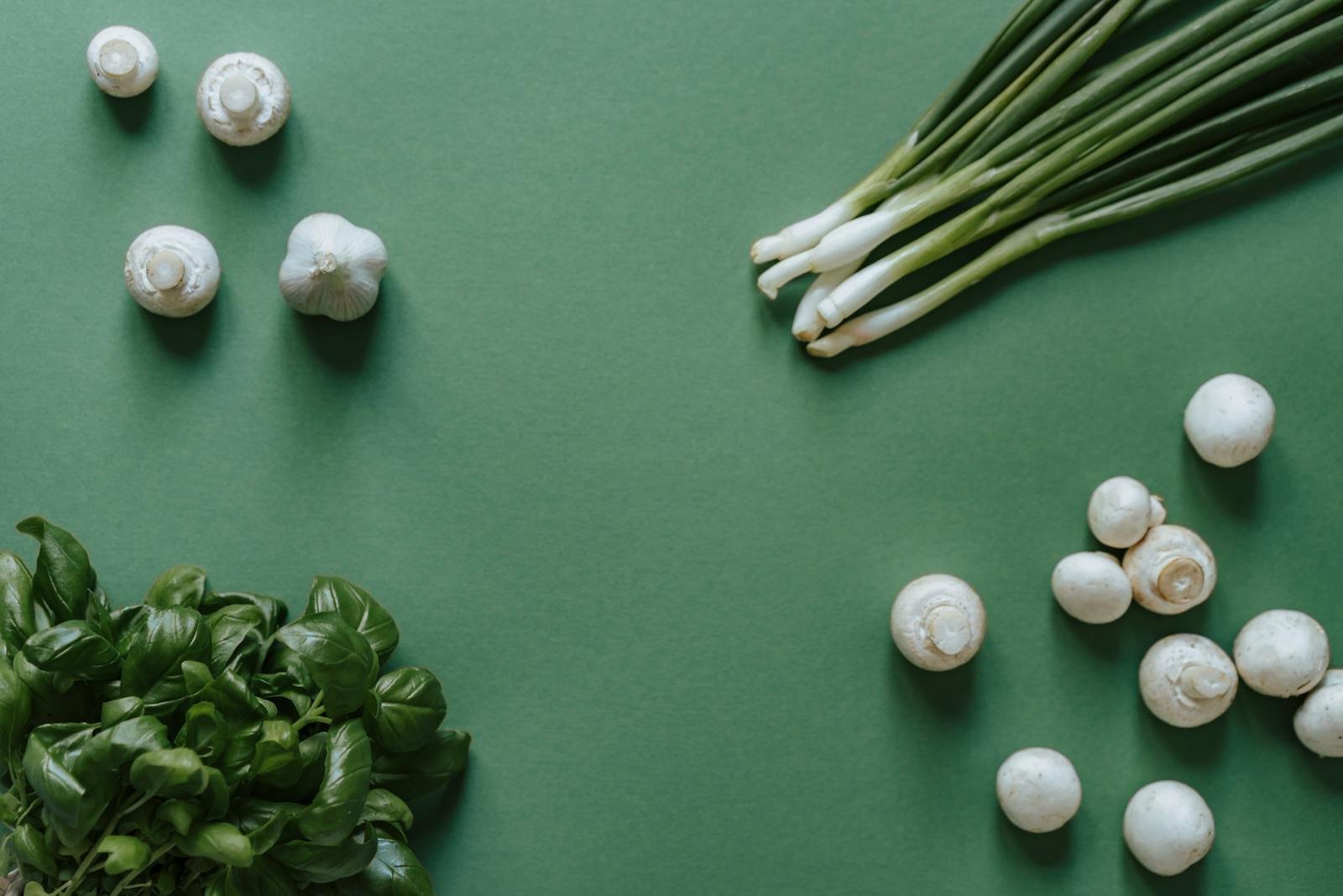 Top-down view of fresh ingredients including basil, mushrooms, garlic, and green onions on a green background.