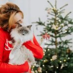A woman embraces a fluffy cat beside a decorated Christmas tree indoors.