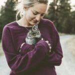 Young woman in a purple sweater smiling down at a kitten she's holding outdoors on a pathway.