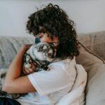 Side view of calm young female owner with dark curly hair and charcoal mask on nose relaxing on soft couch and embracing adorable cat with closed eyes