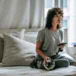 A young woman with curly hair relaxes indoors with her cat, using a smartphone on a cozy bed.