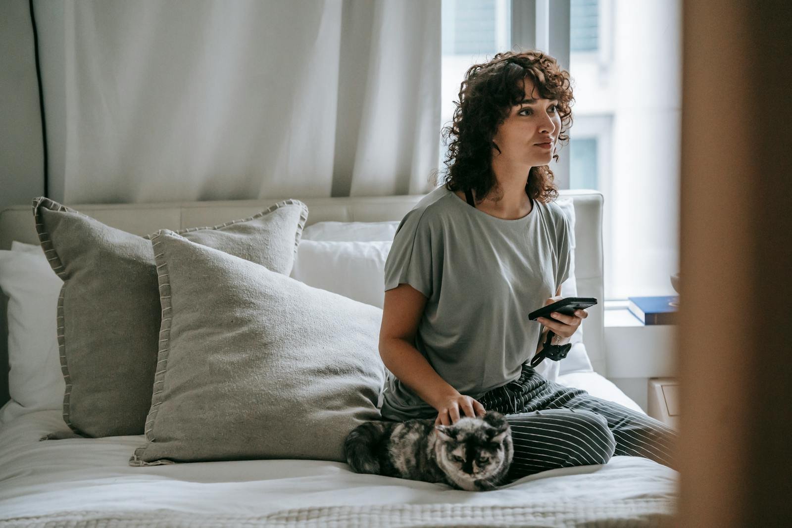 A young woman with curly hair relaxes indoors with her cat, using a smartphone on a cozy bed.