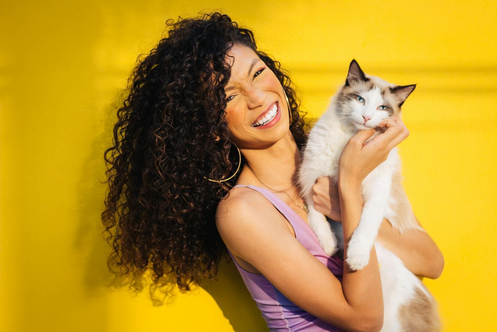 A cheerful woman with curly hair holding a white cat against a vibrant yellow backdrop, exuding happiness.