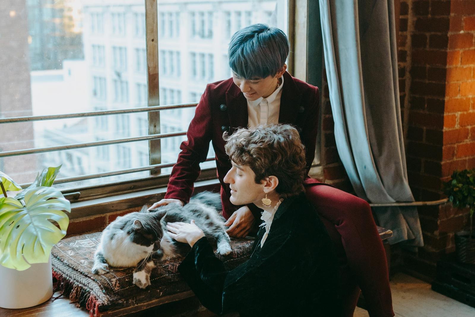 A couple shares a cozy moment with their cat by a large window in an urban apartment.