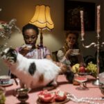 A cozy tea party setting with two women enjoying tea amid candlelight and cats on the dining table.
