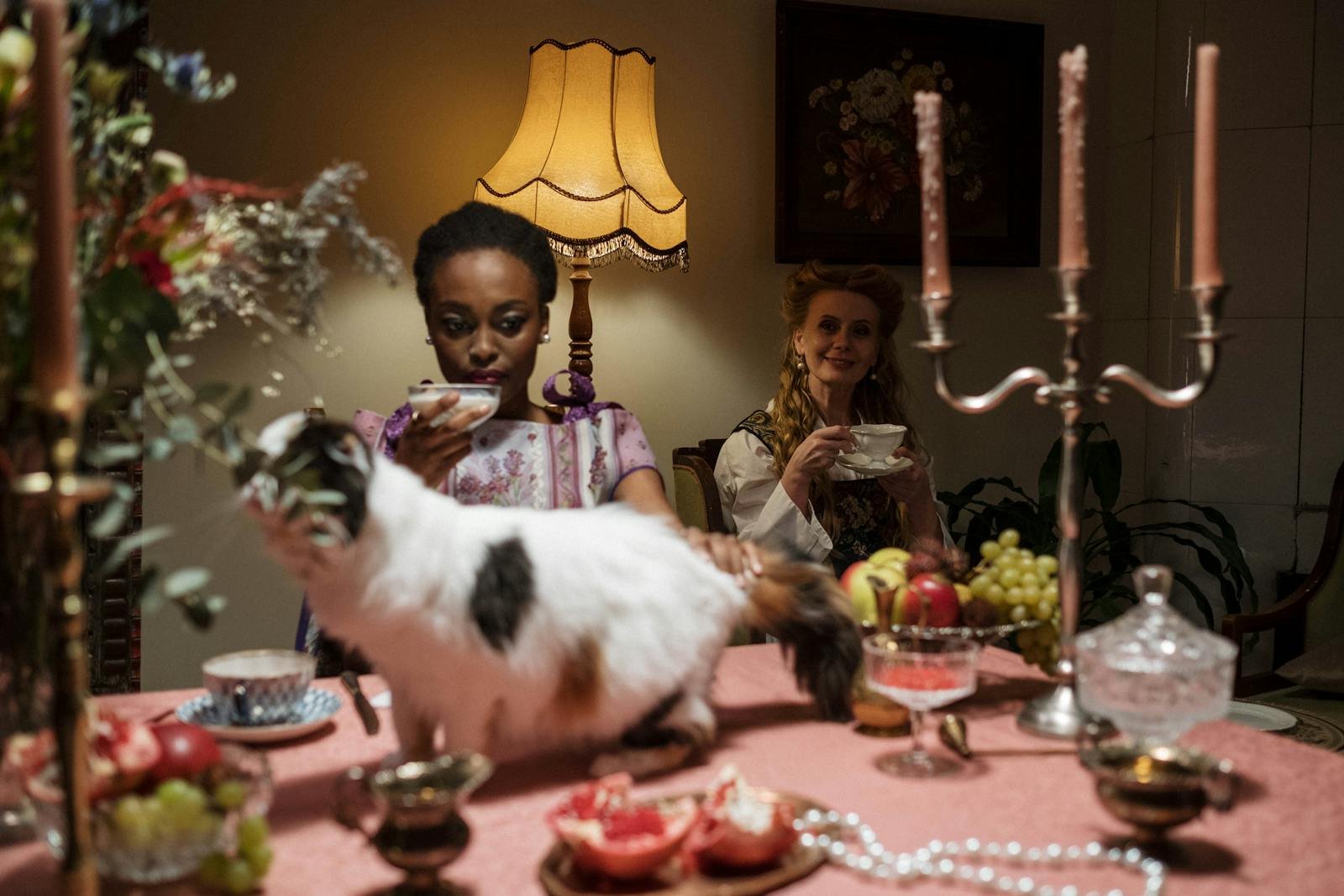 A cozy tea party setting with two women enjoying tea amid candlelight and cats on the dining table.