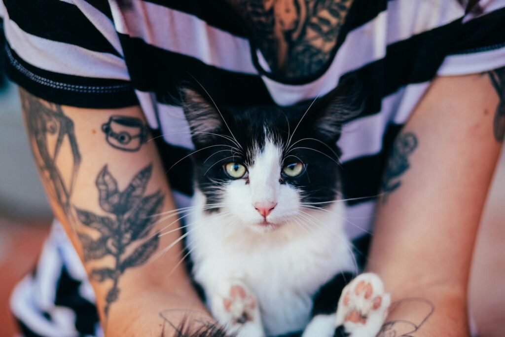 A close-up of a tattooed individual holding a black and white cat, both looking directly at the camera.