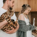 A man holding a Bengal cat while a woman slices fruits in a cozy kitchen setting.