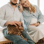 A loving pregnant couple sits by a window with their pet cat, enjoying a serene moment together.