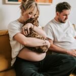 Cozy scene of a pregnant woman with a cat, while her partner works on a laptop on the sofa.
