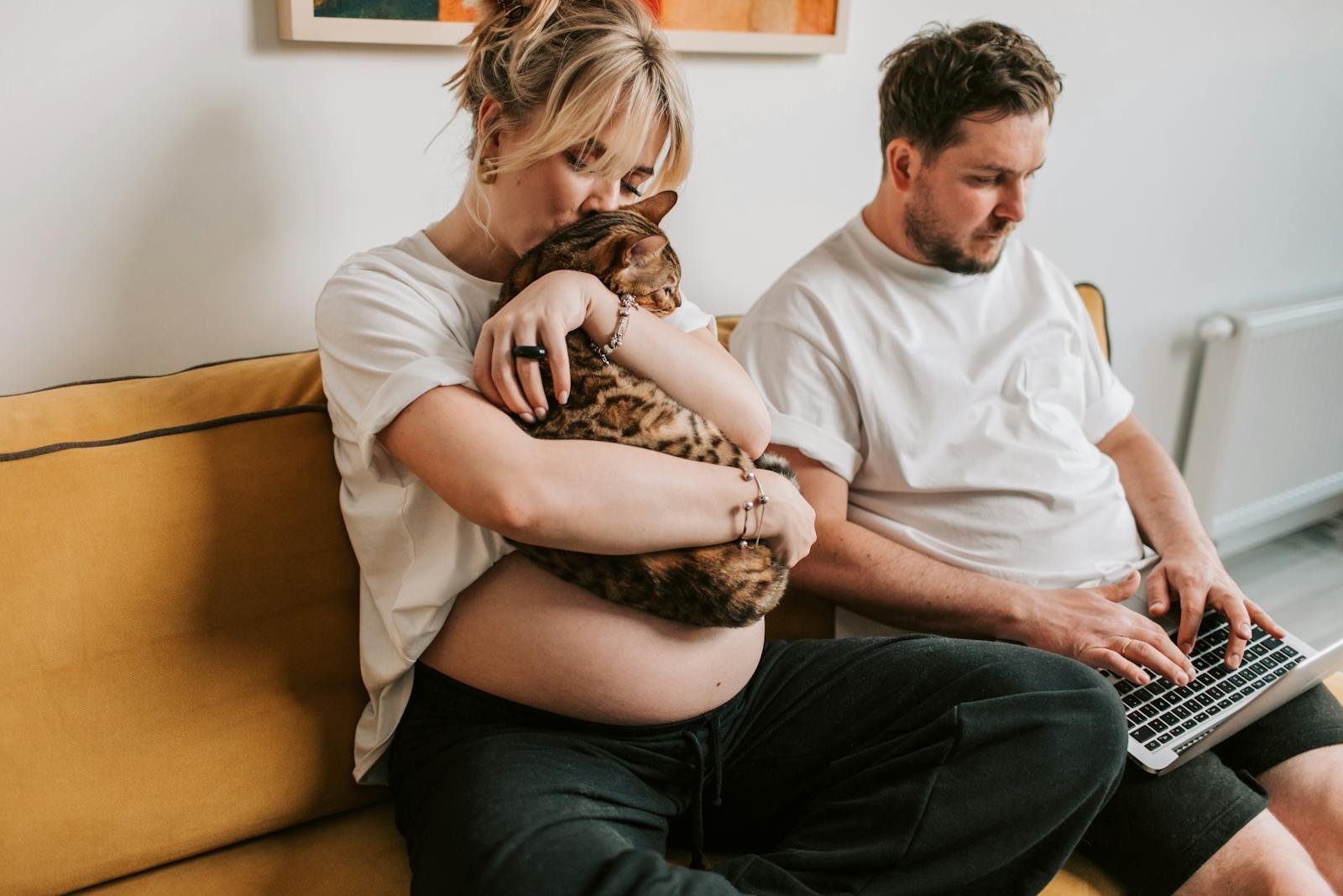 Cozy scene of a pregnant woman with a cat, while her partner works on a laptop on the sofa.