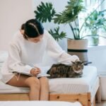 An Asian woman in a white outfit journaling while petting her cat on a bed surrounded by plants.