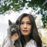 Portrait of a young woman holding a gray cat outdoors on a sunny day.
