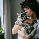A woman lovingly cuddles her gray cat by the window, capturing a serene moment indoors.