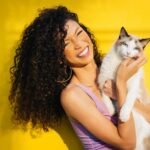 A cheerful woman with curly hair holding a white cat against a vibrant yellow backdrop, exuding happiness.