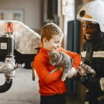 A firefighter assists a child in holding a kitten inside a fire station.