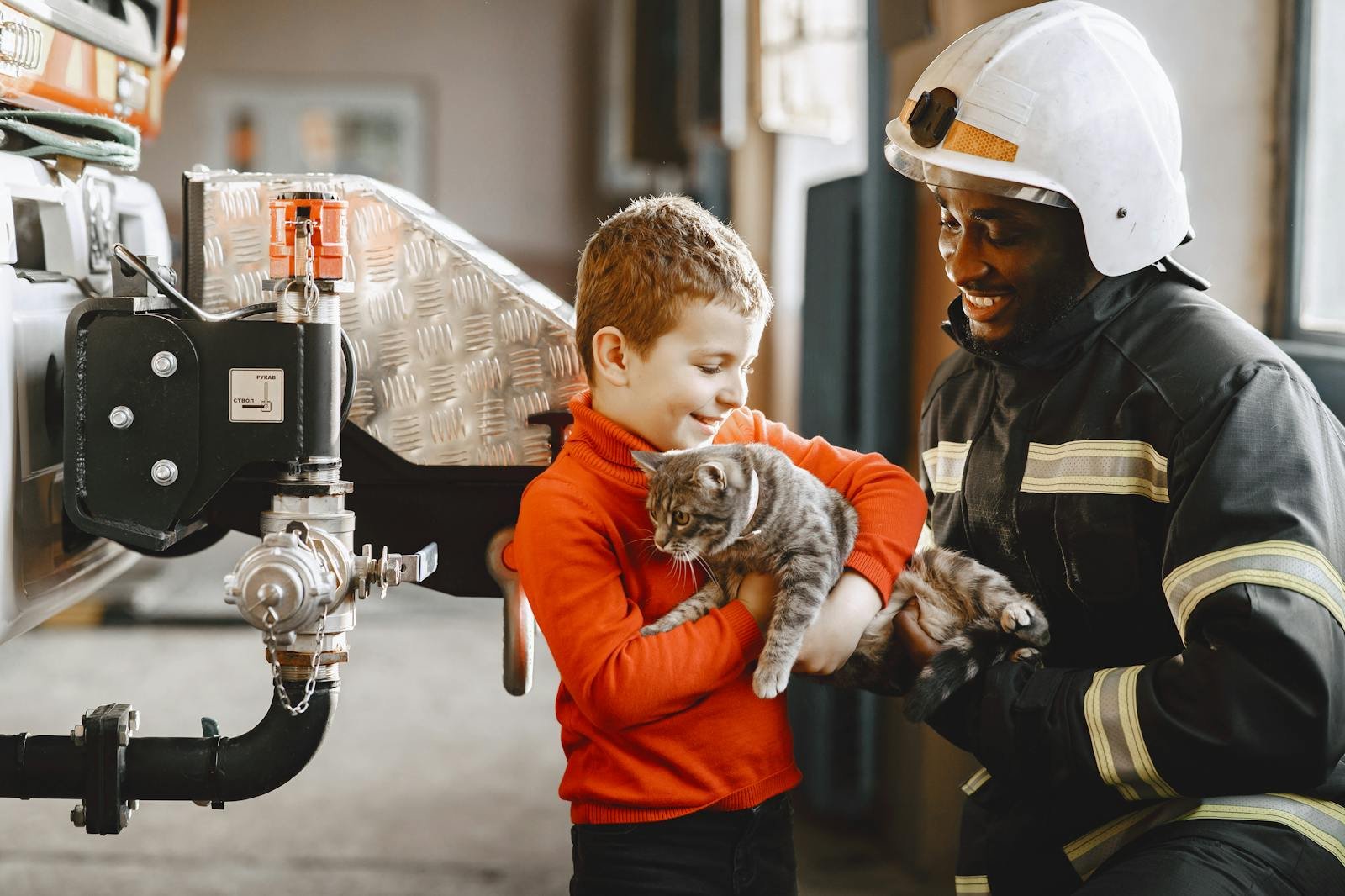 A firefighter assists a child in holding a kitten inside a fire station.
