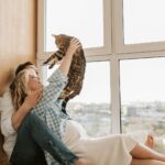 A joyful pregnant couple relaxing by a window, enjoying time with their pet cat.