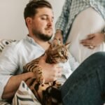 A man sits comfortably holding a Bengal cat while his pregnant partner stands close, creating a cozy indoor scene.