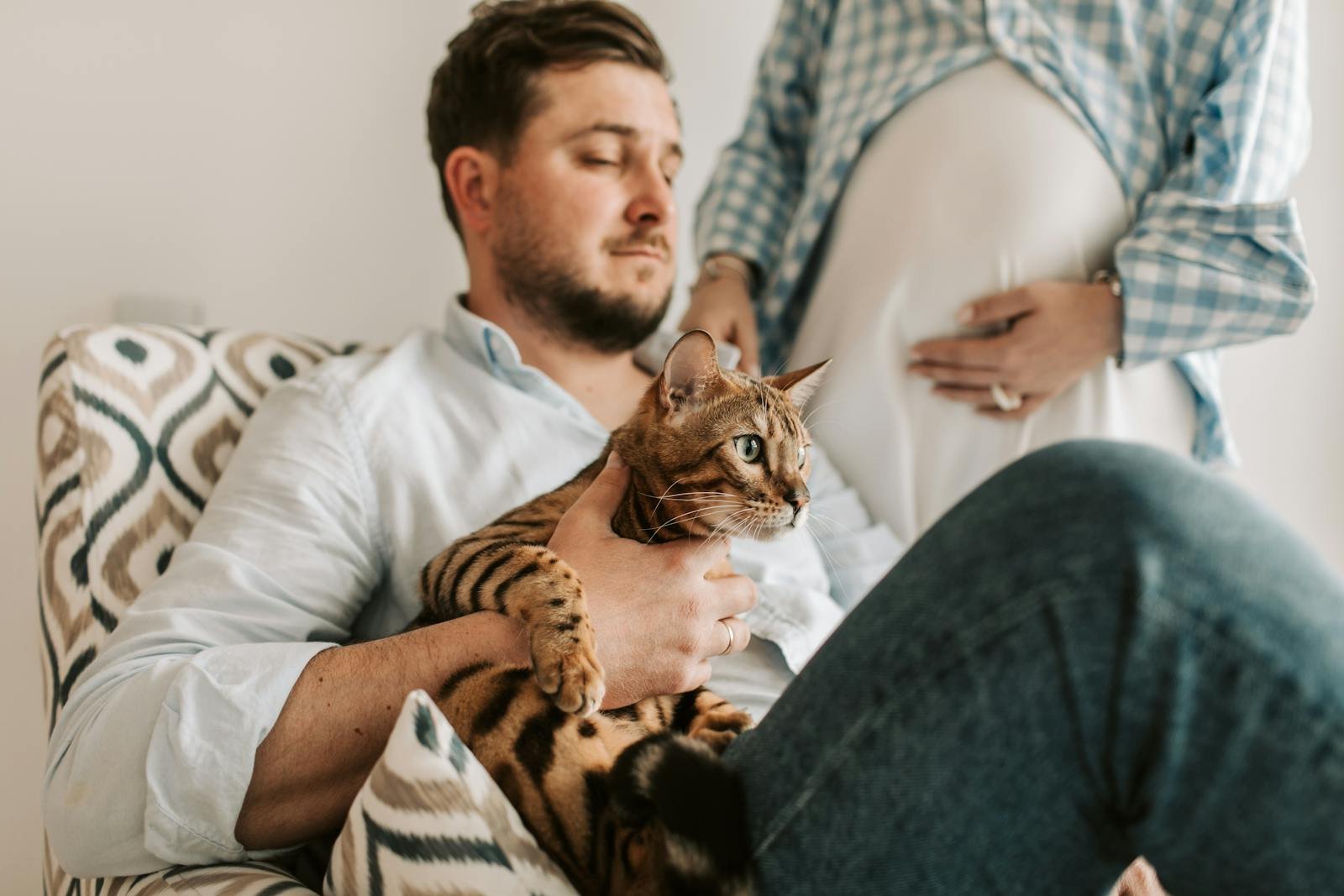 A man sits comfortably holding a Bengal cat while his pregnant partner stands close, creating a cozy indoor scene.