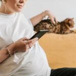 A pregnant woman sitting indoors, using a smartphone, with a cat on the sofa.