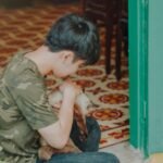 A young boy in a camouflage shirt lovingly cuddles a cat near a green doorway indoors.