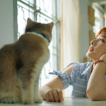 An Asian woman gazes thoughtfully beside a curious cat indoors.