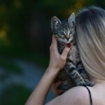 A young woman with blonde hair holds a tabby kitten outdoors, creating a tender moment.