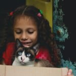A young girl with curly hair smiles while holding a cat in a cardboard box.