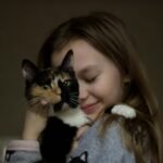 A young girl lovingly hugs a calico cat indoors, showing affection and friendship.
