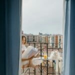 A woman enjoys reading on her balcony with a white cat and a glass of juice.