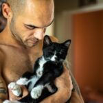Shirtless man holding a tuxedo cat affectionately. Indoor setting, warm tones.