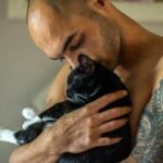 Shirtless man lovingly holding a black cat indoors. Tattoo visible.